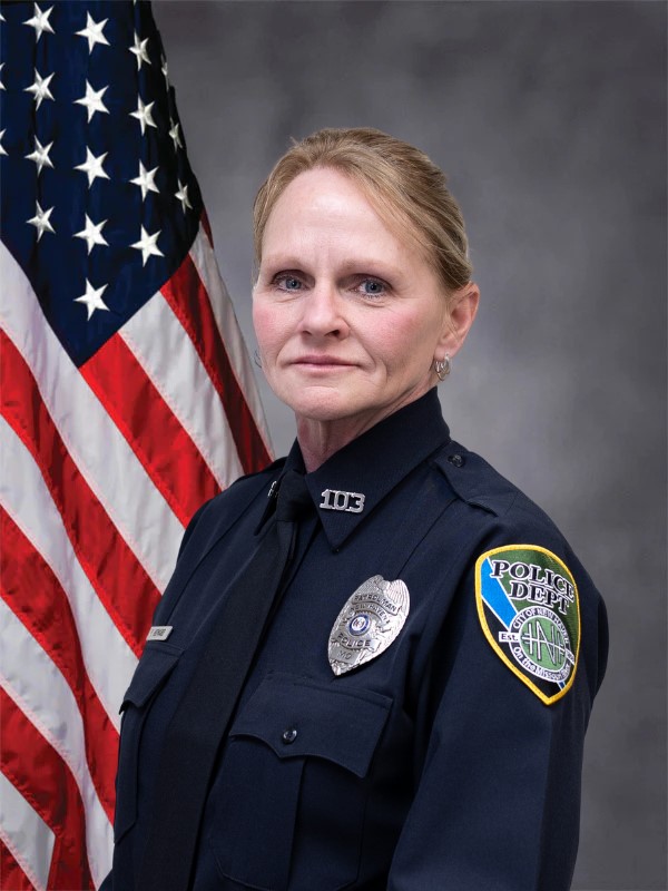 Senior police officer portrait in dark uniform with badge and “103” nameplate; U.S. flag behind her.